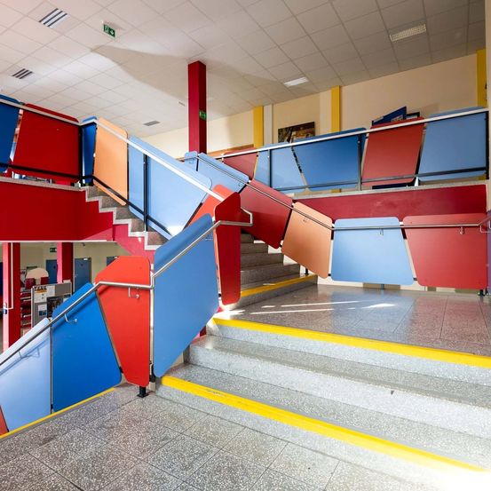 Mehrfarbige Treppe mit Handläufen in einem Gebäudeinneren. Die Treppe besteht aus verschiedenen Farben und Formen.