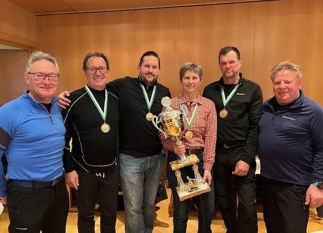 Eine Gruppe von Menschen steht in einem Raum, alle tragen Medaillen, lächeln und posieren für ein Foto. Sie halten eine Trophäe.