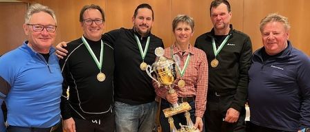 Eine Gruppe von Menschen steht in einem Raum, alle tragen Medaillen, lächeln und posieren für ein Foto. Sie halten eine Trophäe.