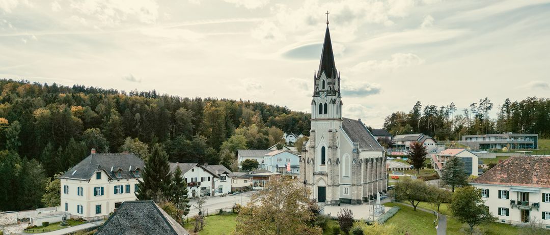 Ein Luftbild einer Kirche mit einem Kirchturm, umgeben von Bäumen und Häusern unter einem bewölkten Himmel.
