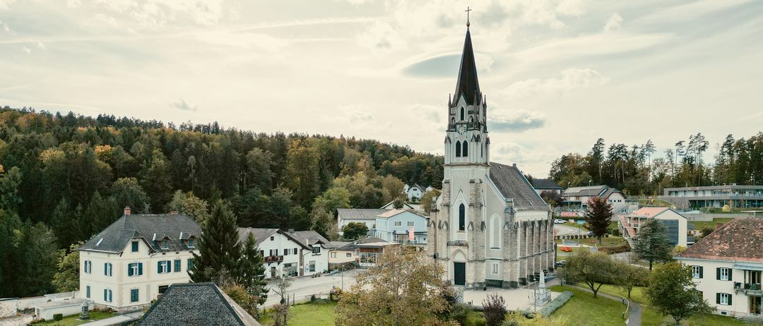 Luftaufnahme einer kleinen Stadt mit einer Kirche, umgeben von Bäumen und Häusern. Die Kirche hat einen Turm und befindet sich im Zentrum der Stadt.