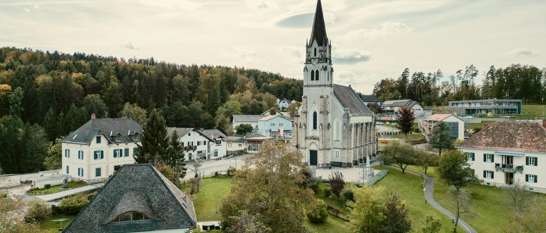 Eine friedliche Luftaufnahme eines Dorfes mit einem hohen Kirchturm, umgeben von üppigen Bäumen und Häusern, unter einem bewölkten Himmel.