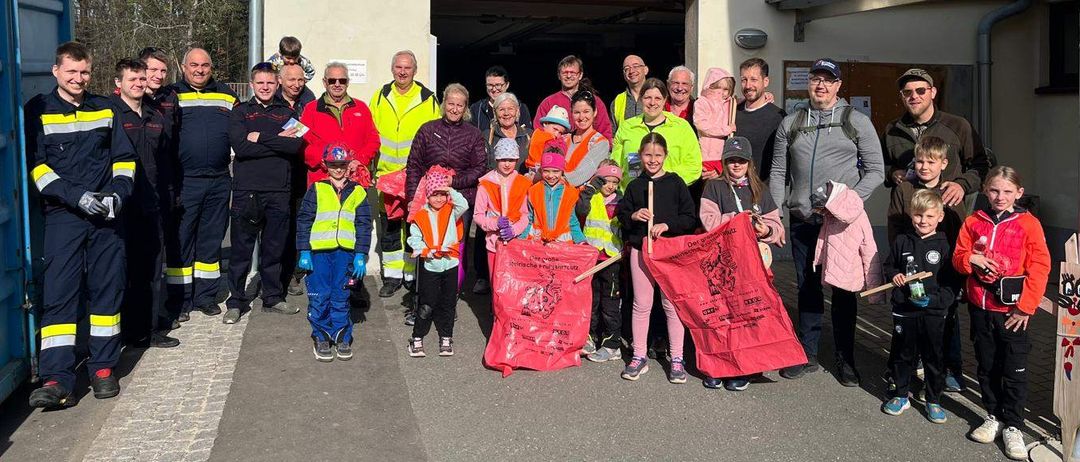 Eine Gruppe von Menschen posiert für ein Foto vor einem Gebäude mit der Aufschrift 'Altstoffsammlungszentrum'. Einige halten rote Banner. Dazu gehören Feuerwehrleute, Kinder und Erwachsene, alle warm gekleidet. Bäume sind im Hintergrund zu sehen.