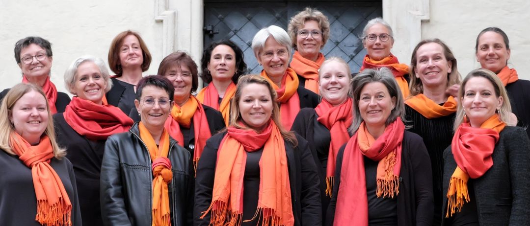 Ein Chor von Frauen steht auf Stufen und trägt schwarze und orange Schals, lächelt für ein Gruppenfoto vor einem Gebäude.