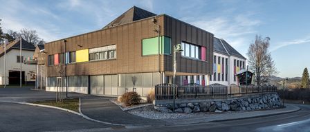 A modern building with a brown wooden exterior and colorful windows is in a corner of the road, surrounded by a stone barrier and a small tree.