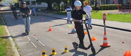 A group of people are riding scooters on a street, navigating through cones. A person in the foreground is wearing a helmet and sneakers. In the background, there is a building with a stop sign.
