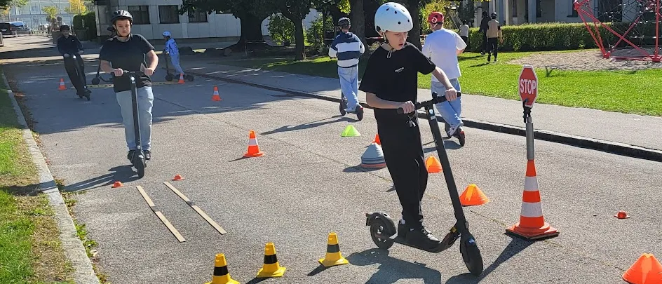 Eine Gruppe von Menschen fährt auf einer Straße mit Rollern und navigiert durch Hütchen. Eine Person im Vordergrund trägt einen Helm und Turnschuhe. Im Hintergrund befindet sich ein Gebäude mit einem Stoppschild.