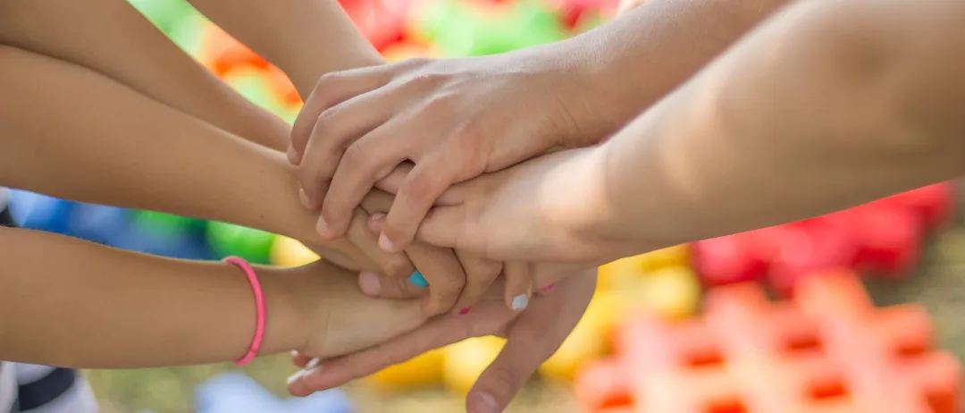 Nahaufnahme mehrerer Hände von Kindern, die sich gegenseitig in einem Kreis halten, mit bunten Spielzeugen im Hintergrund.