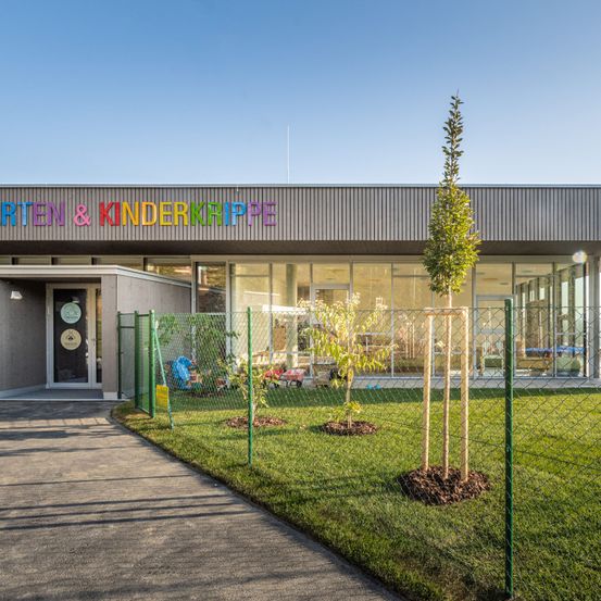 Die Vorderseite eines modernen Gebäudes mit der Aufschrift 'Kindergarten & Kinderkrippe', einem eingezäunten Hof, einem Weg und mehreren jungen Bäumen.