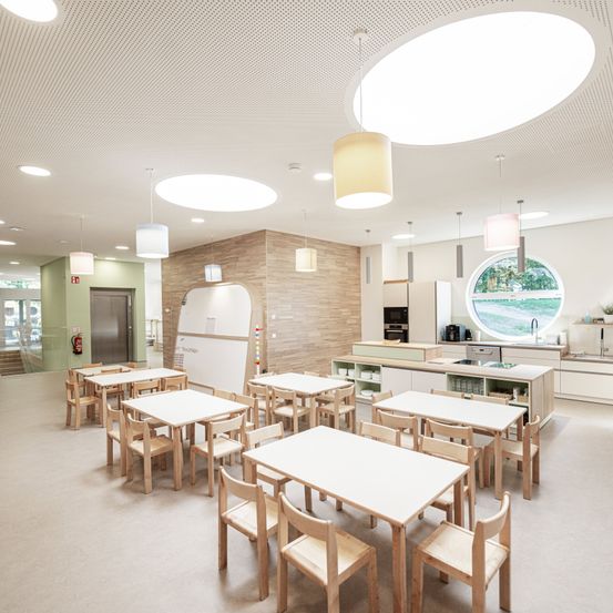 Ein moderner Essbereich mit Holztischen und Stühlen, großen runden Leuchten und einem runden Fenster. Küchengeräte und ein Waschbecken sind im Hintergrund zu sehen.