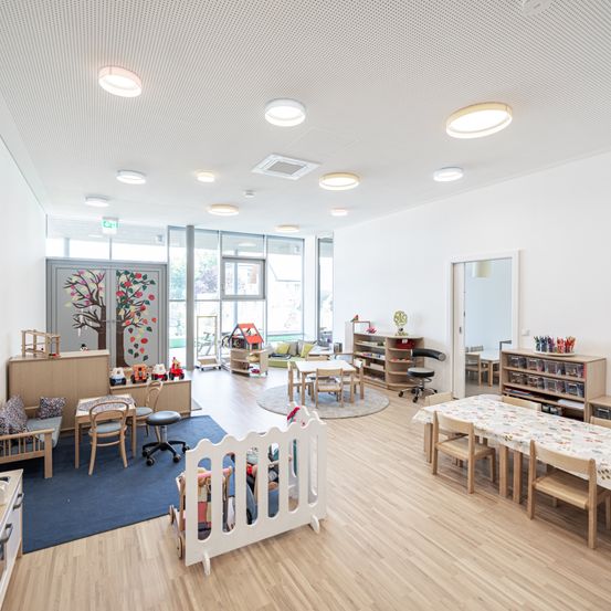 Ein Innenansicht eines Kinderzimmers mit weißen Wänden, Holzböden und verschiedenen Möbeln. Das Zimmer verfügt über Tische, Stühle, einen blauen Teppich und eine Babywiege. Es gibt Regale mit Spielzeug und Büchern sowie eine Glastür, die zu einem Flur führt. Die Decke hat Einbauleuchten.