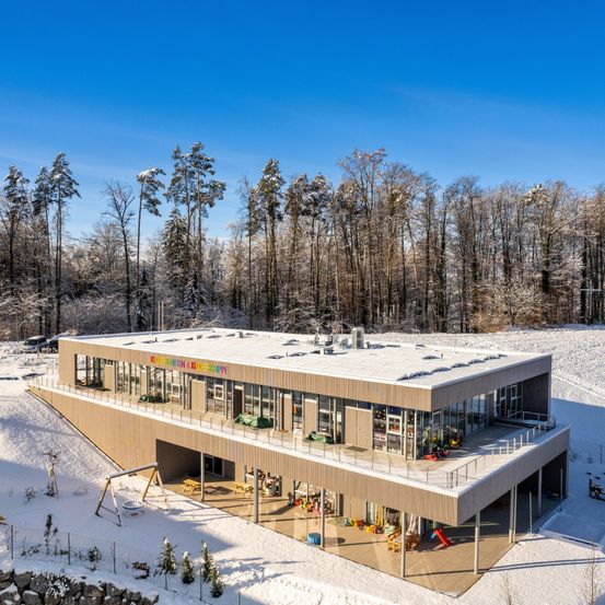 Ein modernes Gebäude mit flachem Dach, umgeben von Schnee und Bäumen im Winter. Das Gebäude hat Glasfenster und einen Spielplatz davor.