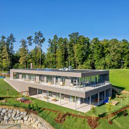 Eine Luftaufnahme eines modernen, mehrstöckigen Gebäudes mit Glaswänden und einem Solardach, eingebettet in eine üppige grüne Landschaft mit Spielplatz und Picknickbereich im Vordergrund.