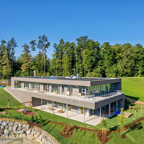 Eine Luftaufnahme eines modernen, mehrstöckigen Gebäudes mit Glaswänden und einem Solardach, eingebettet in eine üppige grüne Landschaft mit Spielplatz und Picknickbereich im Vordergrund.