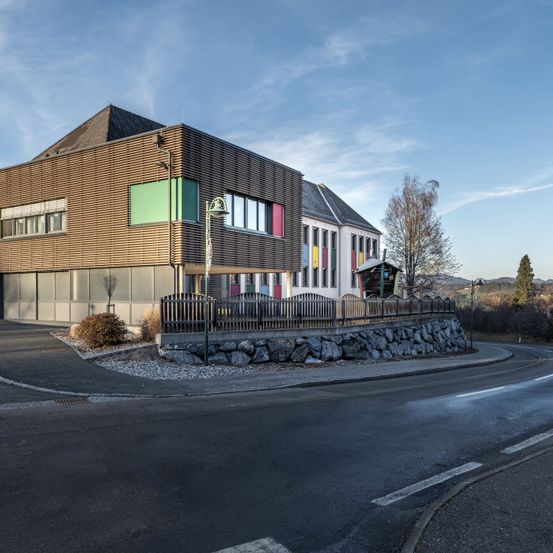 Ein modernes Gebäude mit brauner Fassade und farbenfrohen Fenstern steht am Straßenrand mit einem Laternenpfosten und Zaun. Im Hintergrund befinden sich Bäume und ein klarer Himmel.