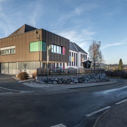 Ein modernes Gebäude mit brauner Fassade und farbenfrohen Fenstern steht am Straßenrand mit einem Laternenpfosten und Zaun. Im Hintergrund befinden sich Bäume und ein klarer Himmel.