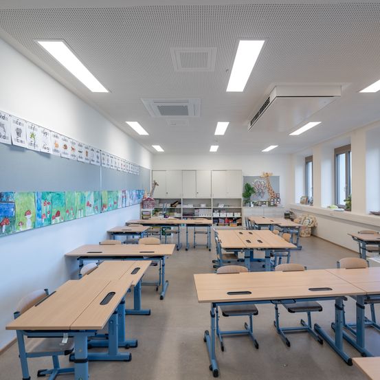 Ein leerer Klassenraum mit mehreren Tischen und Stühlen in Reihen angeordnet. Die Wände sind weiß gestrichen, und es gibt farbenfrohe Poster an einer Wand. Die Decke hat mehrere Lichter.