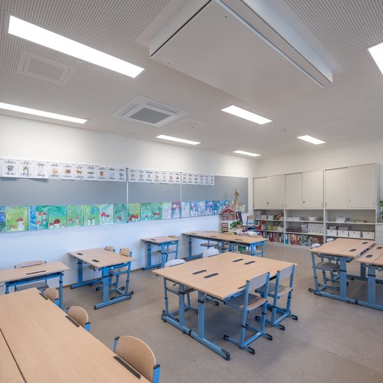 Ein leerer Klassenraum mit blauen Tischen und Stühlen, weißen Wänden und einer Decke mit Lichtern. Bunte Gemälde schmücken die Wand, und es gibt einen Schrank mit Büchern.