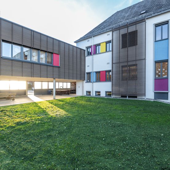 Ein modernes Gebäude mit Glasfenstern, farbigen Paneelen und einem gepflegten Rasen. Die Sonne scheint hell und wirft Schatten auf das Gras.