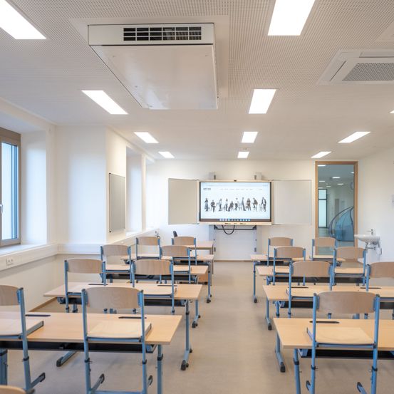 Ein leerer Klassenraum mit Reihen von Tischen und Stühlen, einer Projektionsleinwand an der Wand und einer Tafel. Der Raum ist gut beleuchtet mit Deckenlampen.