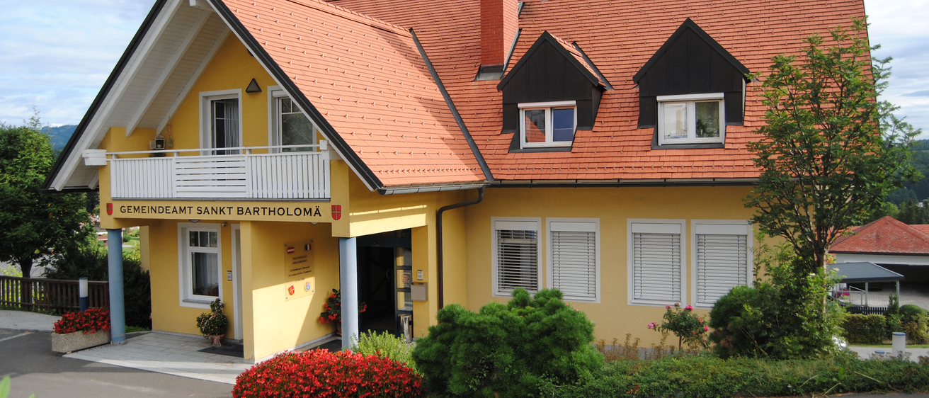 Ein gelbes Haus mit rotem Dach und schwarzem Rand. Der Name 'Deamt Sankt Bartholoma' steht über dem Eingang. Vor dem Haus stehen Pflanzen und Blumen.