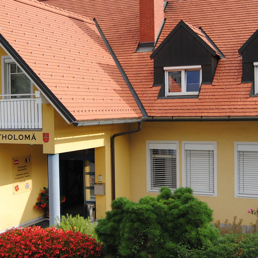 Ein gelbes Haus mit rotem Dach und schwarzem Rand. Der Name 'Deamt Sankt Bartholoma' steht über dem Eingang. Vor dem Haus stehen Pflanzen und Blumen.
