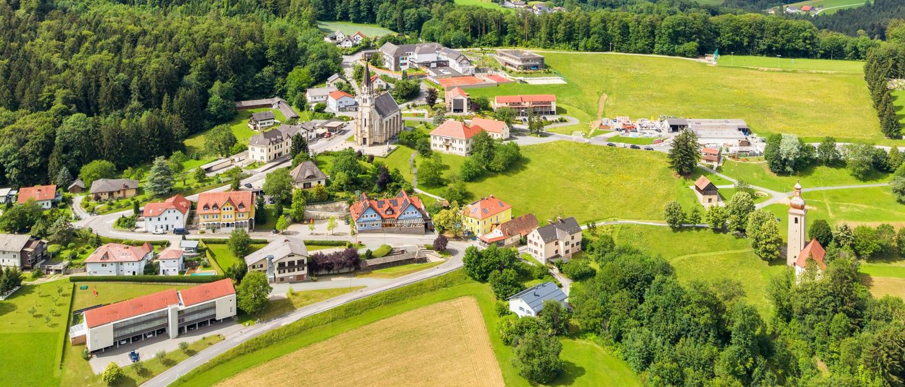 Luftaufnahme eines kleinen Dorfes umgeben von üppigen grünen Bäumen, Häusern und Bergen in der Ferne.
