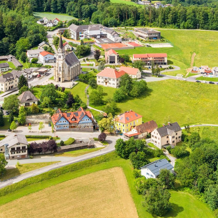 Luftaufnahme eines kleinen Dorfes umgeben von üppigen grünen Bäumen, Häusern und Bergen in der Ferne.