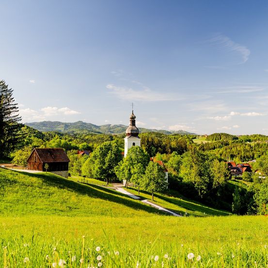 Ein kleines Holzhaus und ein Kirchturm sind von üppigen grünen Hügeln umgeben, unter einem klaren blauen Himmel.