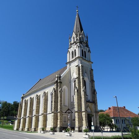 Eine Kirche mit einem Turm und einer Statue davor befindet sich in einer ländlichen Gegend unter einem klaren blauen Himmel.