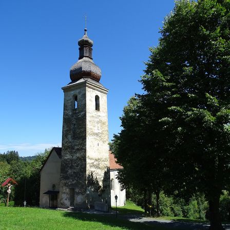 Eine alte Kirche mit einem hohen Turm und einem kuppelförmigen Dach ist von saftigem grünem Gras und Bäumen umgeben unter einem klaren blauen Himmel.