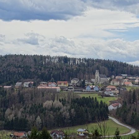 Eine Stadt in den Bergen mit einer Mischung aus Häusern, einer Kirche und einer kurvenreichen Straße, umgeben von Bäumen unter einem bewölkten Himmel.