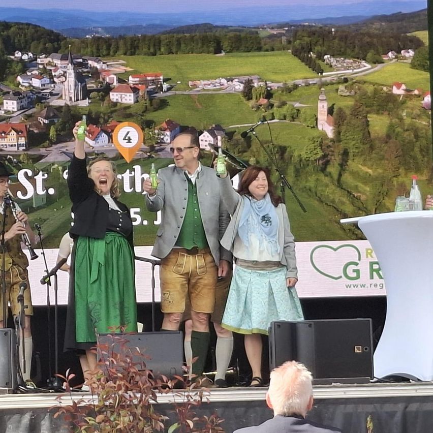 A group of performers stand on a stage, two raising green bottles. A backdrop displays a village with hills in the distance.