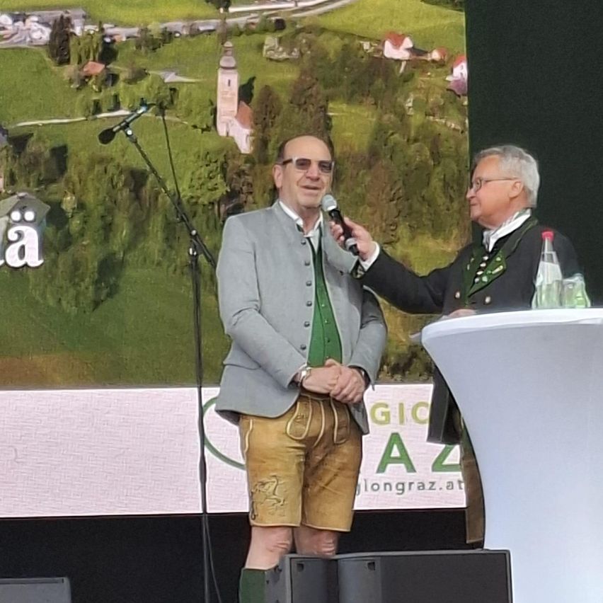 Two men are on stage with a microphone and a podium. One man holds the microphone, and the other man stands beside him. Behind them is a large screen showing a rural landscape.
