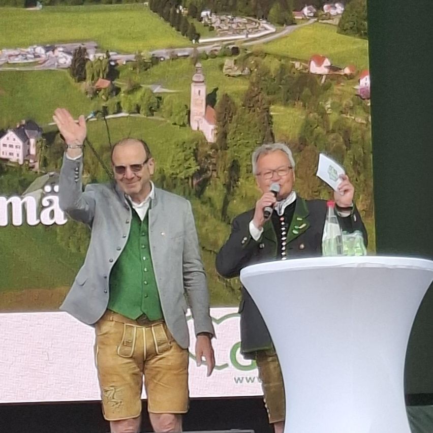 Two men in traditional German attire stand on stage. One waves and smiles, the other holds a microphone and a booklet. A scenic backdrop shows a village.