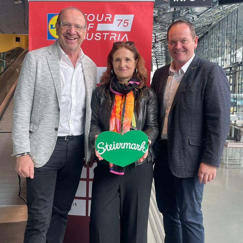 Three people stand in front of a red banner, smiling. The woman holds a green heart. They appear to be posing for a photo. Behind them, a metal structure with a conveyor belt is visible.