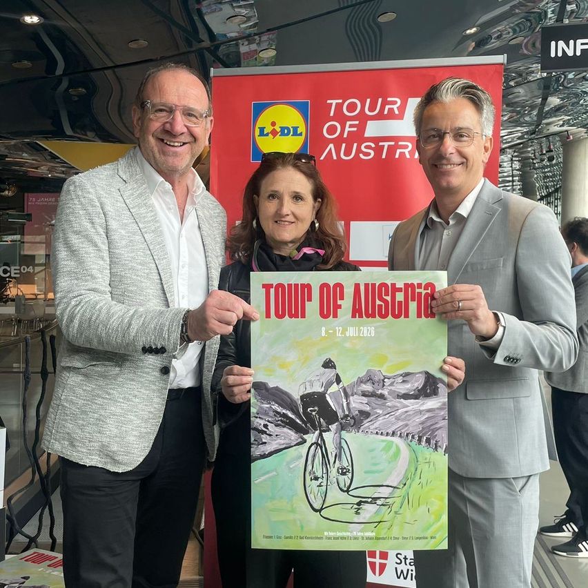 Three people stand in front of a poster, two men and one woman, all wearing formal attire. The poster features a cyclist on a mountain road, with 'Tour of Austria' written above the cyclist.