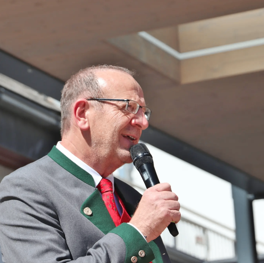Ein Mann in einem formellen Anzug mit grünem Kragen und roter Krawatte, spricht in ein Mikrofon.