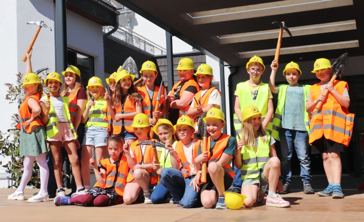 Gruppe von Kindern in Sicherheitswesten und Bauhelmen, die Werkzeuge halten und vor einem Gebäude für ein Foto posieren.