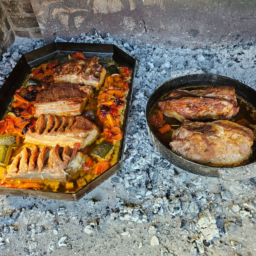 Zwei Kochtöpfe mit gebratenem Fleisch und Gemüse stehen auf einem Grill im Freien. Das Fleisch hat eine knusprige, goldbraune Außenseite. Das Gemüse besteht aus einer Mischung aus Karotten, Paprika und Zucchini.