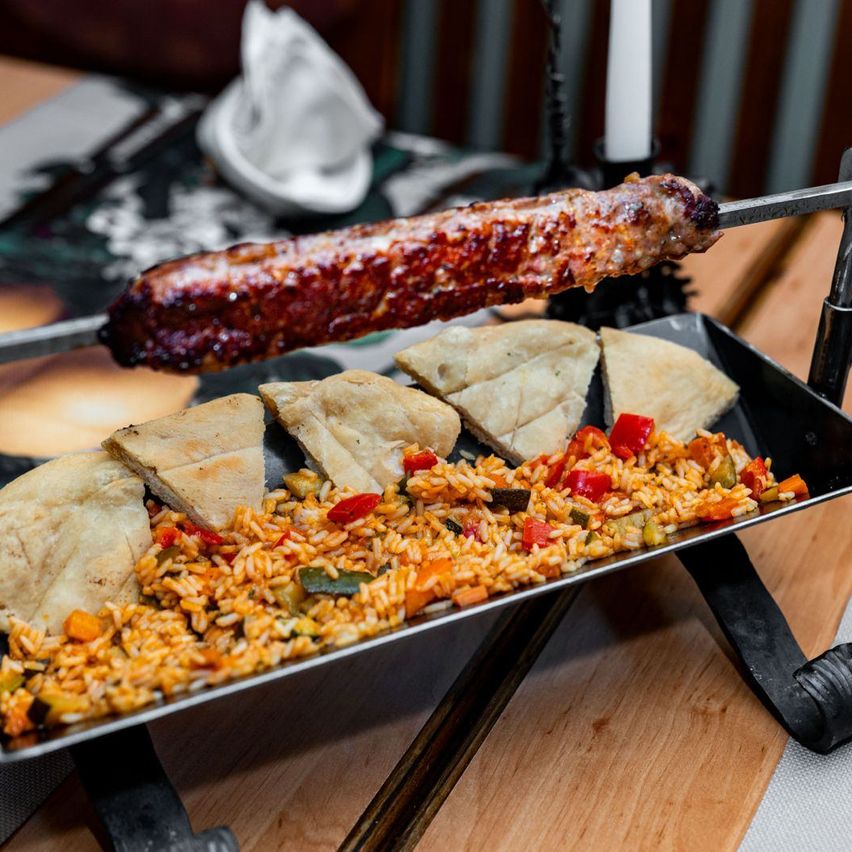 Ein gegrillter Fleischspieß ruht über einem Tablett mit Reis, Gemüse und Fladenbrot, präsentiert auf einem Holztisch mit dekorativen Elementen.