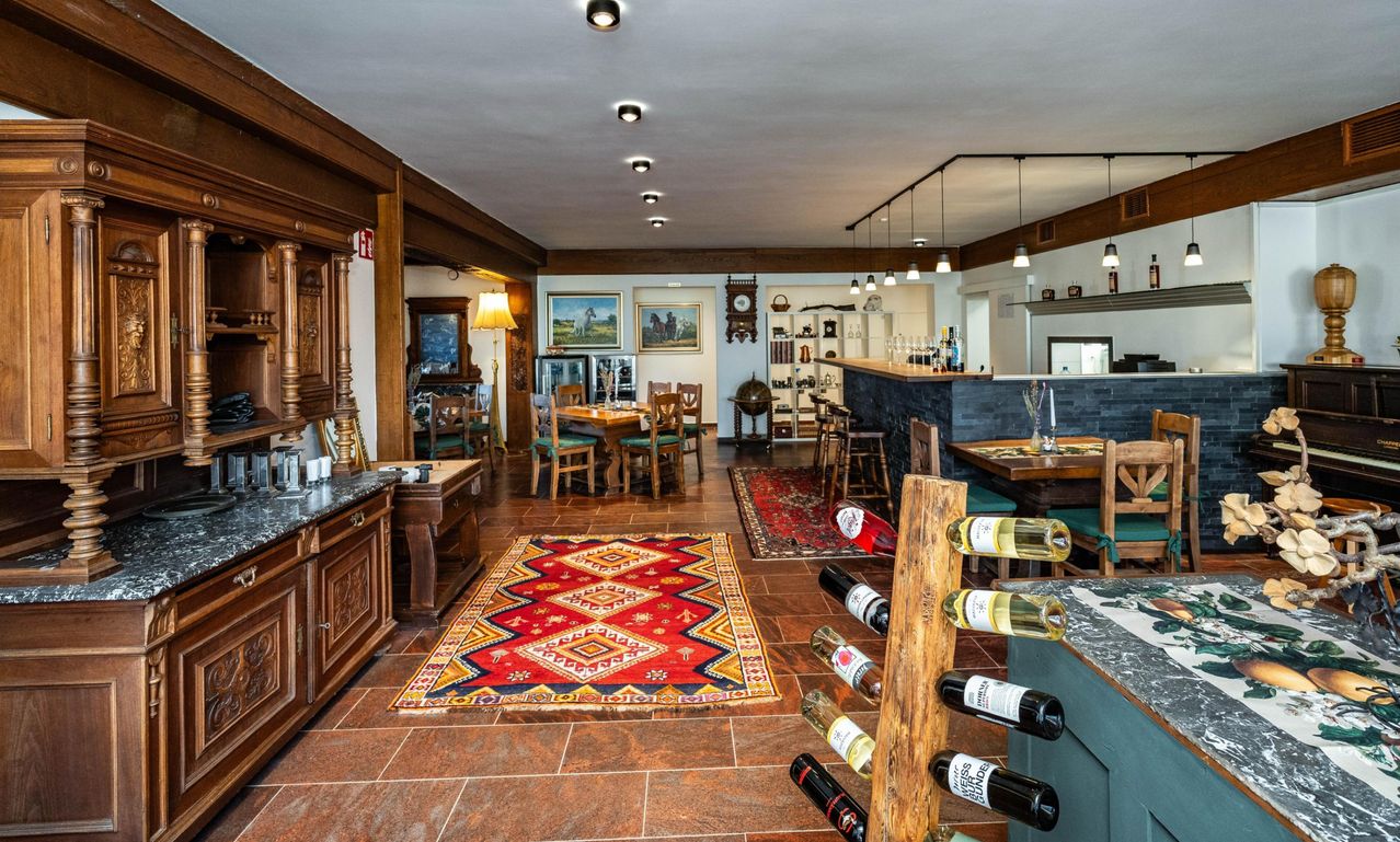 Ein gemütliches Restaurant mit Holzmöbeln, einem Teppich und einem Weinregal. Flaschen sind auf einem Holzständer ausgestellt. Ein Tisch mit Weingläsern und ein Schrank mit Flaschen.