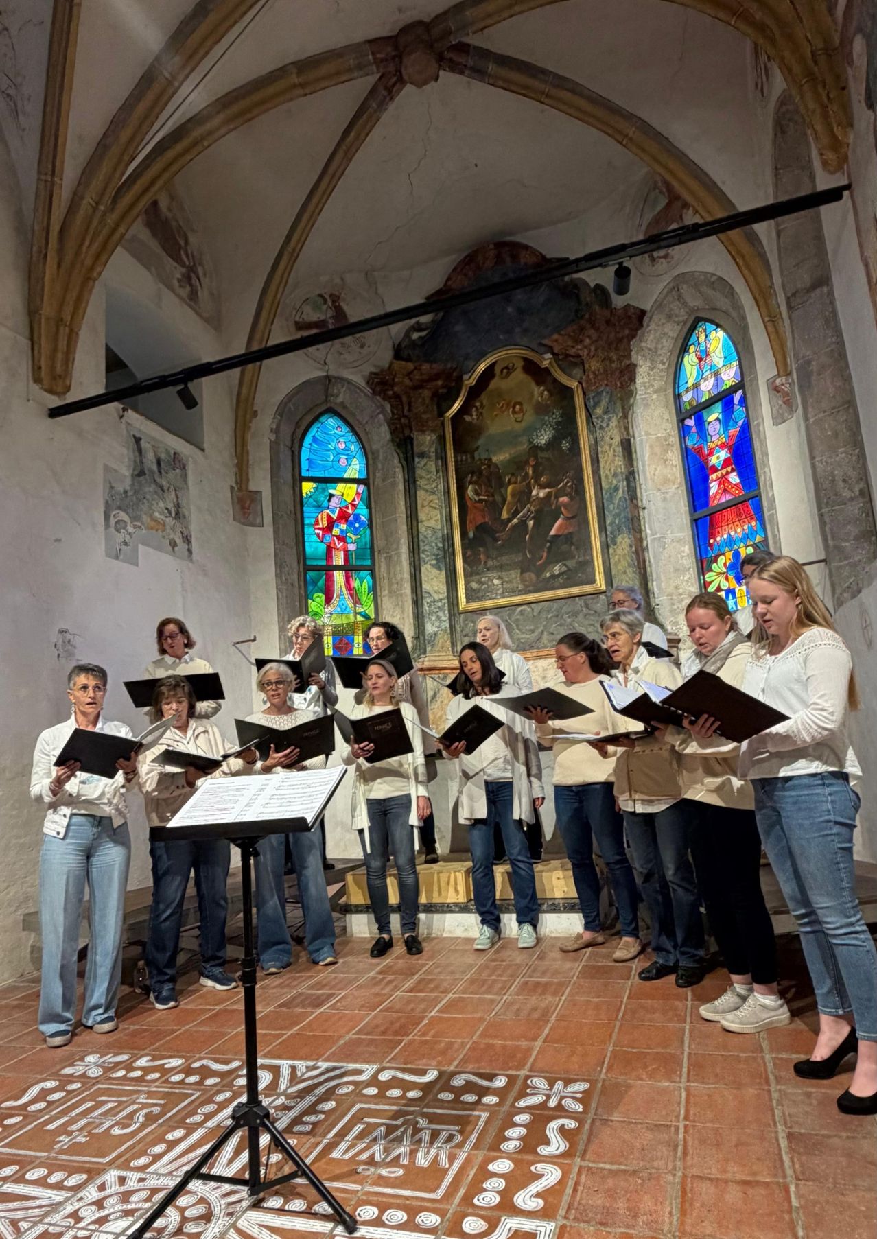 Ein Frauenchor steht in einer Kirche, hält Notenblätter in den Händen, gekleidet in weißen Oberteilen und Jeans. Dahinter ist ein großes Gemälde montiert, und Buntglasfenster befinden sich auf beiden Seiten.