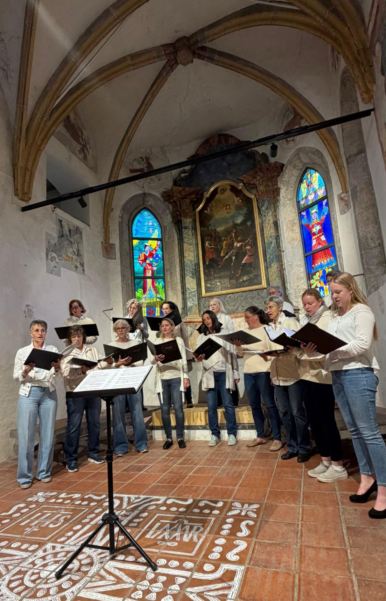 Ein Frauenchor steht in einer Kirche, hält Notenblätter in den Händen, gekleidet in weißen Oberteilen und Jeans. Dahinter ist ein großes Gemälde montiert, und Buntglasfenster befinden sich auf beiden Seiten.