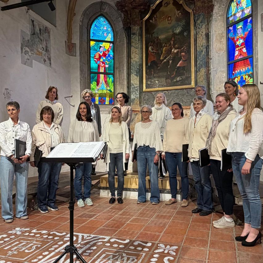 Ein Chor von Frauen in weißen Shirts und Jeans steht in einer Kirche, Musikbücher in der Hand, vor einem Notenständer und Buntglasfenstern.