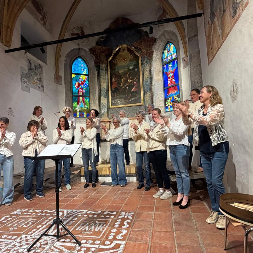 Eine Gruppe von Frauen, gekleidet in weißen Jacken, steht vor einem Notenständer in einer Kapelle. Sie klatschen und scheinen zu feiern. Die Kapelle hat Buntglasfenster und eine Wandmalerei.