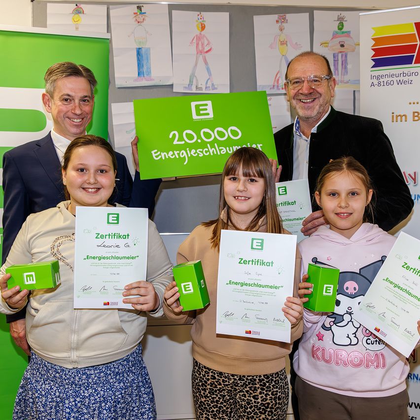 Drei junge Mädchen halten Zertifikate und grüne Boxen in der Hand und stehen vor einem grünen Schild mit '20.000 Energieschlaumeier' darauf, zusammen mit zwei Männern. Sie alle lächeln und posieren für ein Foto. Der Hintergrund ist innen mit grünen Bannern und Zeichnungen an den Wänden.