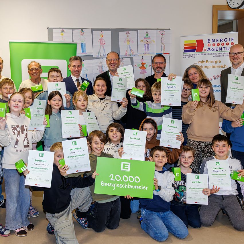 Eine Gruppe von Kindern und Erwachsenen posiert für ein Foto mit grünen Zertifikaten und einem großen grünen Schild, das '20.000 Energieschlauchmeister' besagt. Sie befinden sich in einem Raum mit Postern und einer Uhr an der Wand.