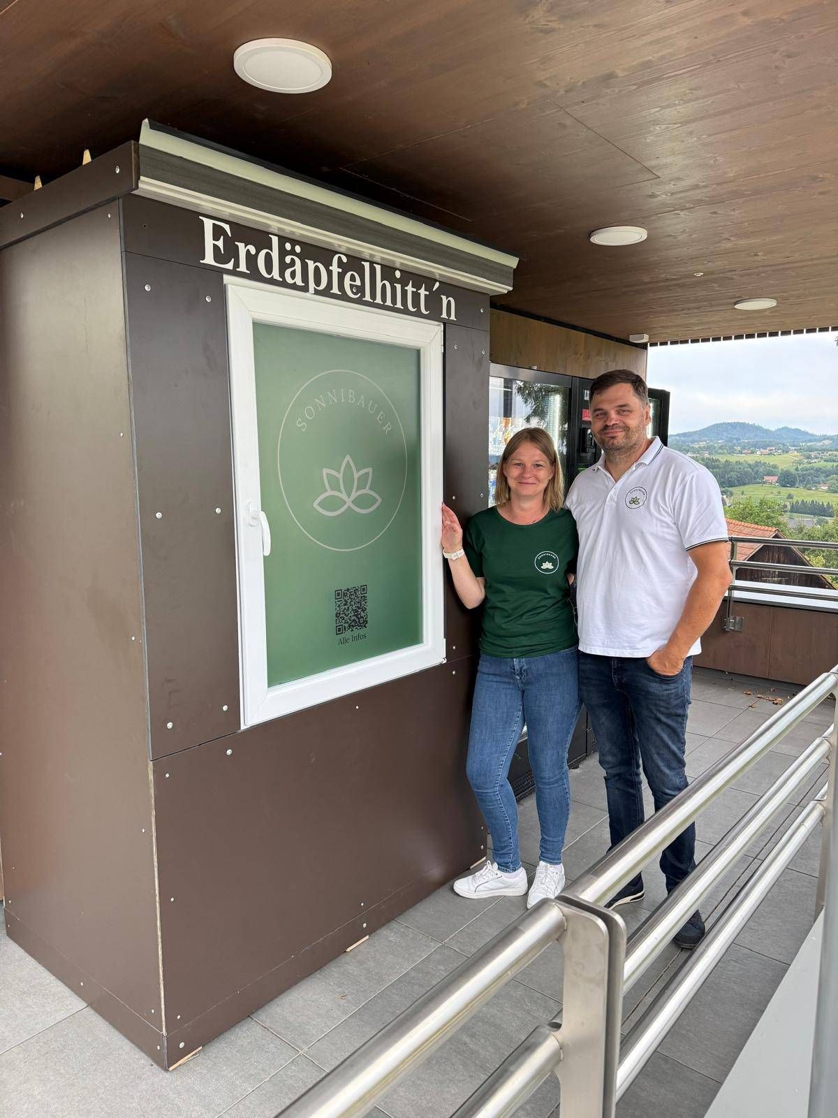 Erdäpfelhütte Sonnbauer mit Betreibern vor dem Selbstbedienungsautomaten in einem ländlichen Gebiet mit Hügeln.