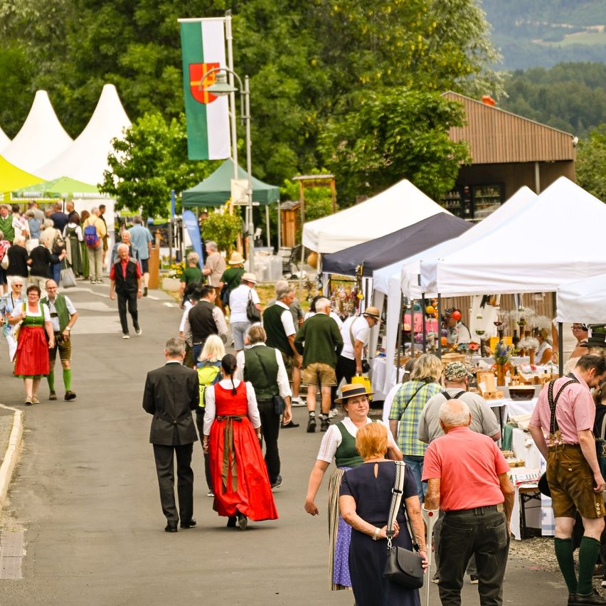 Ein geschäftiger Outdoor-Markt mit vielen Menschen, die gehen, einige in traditioneller deutscher Kleidung. Zelte und Flaggen sind entlang der Straße aufgestellt, und es gibt Bäume und ein Gebäude im Hintergrund.
